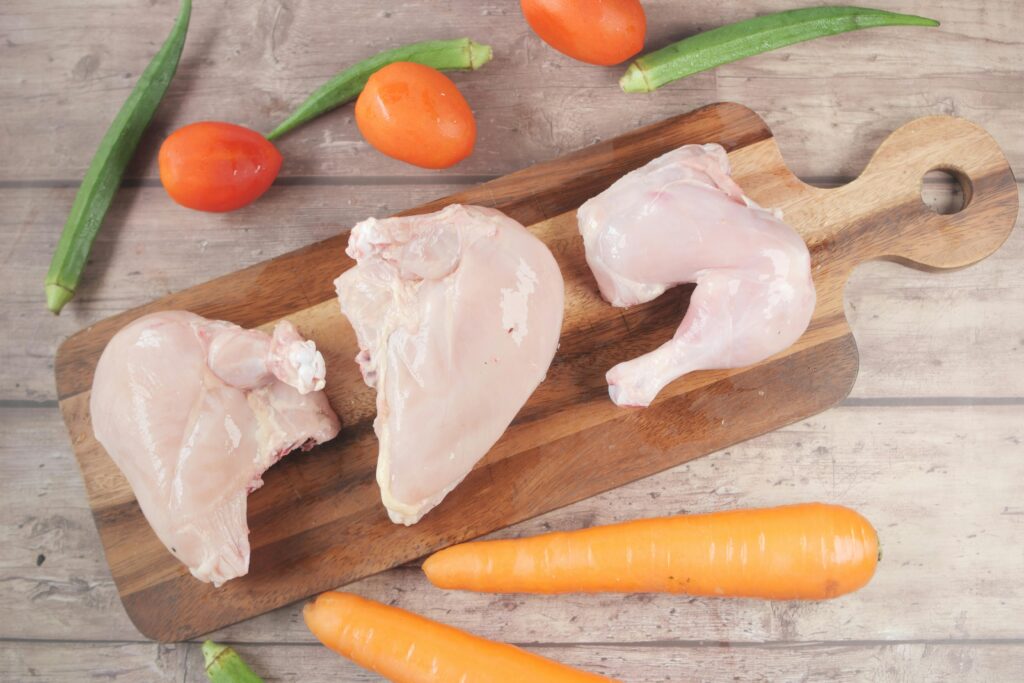 Overhead view of raw chicken, tomatoes, carrots, and okra on a chopping board.