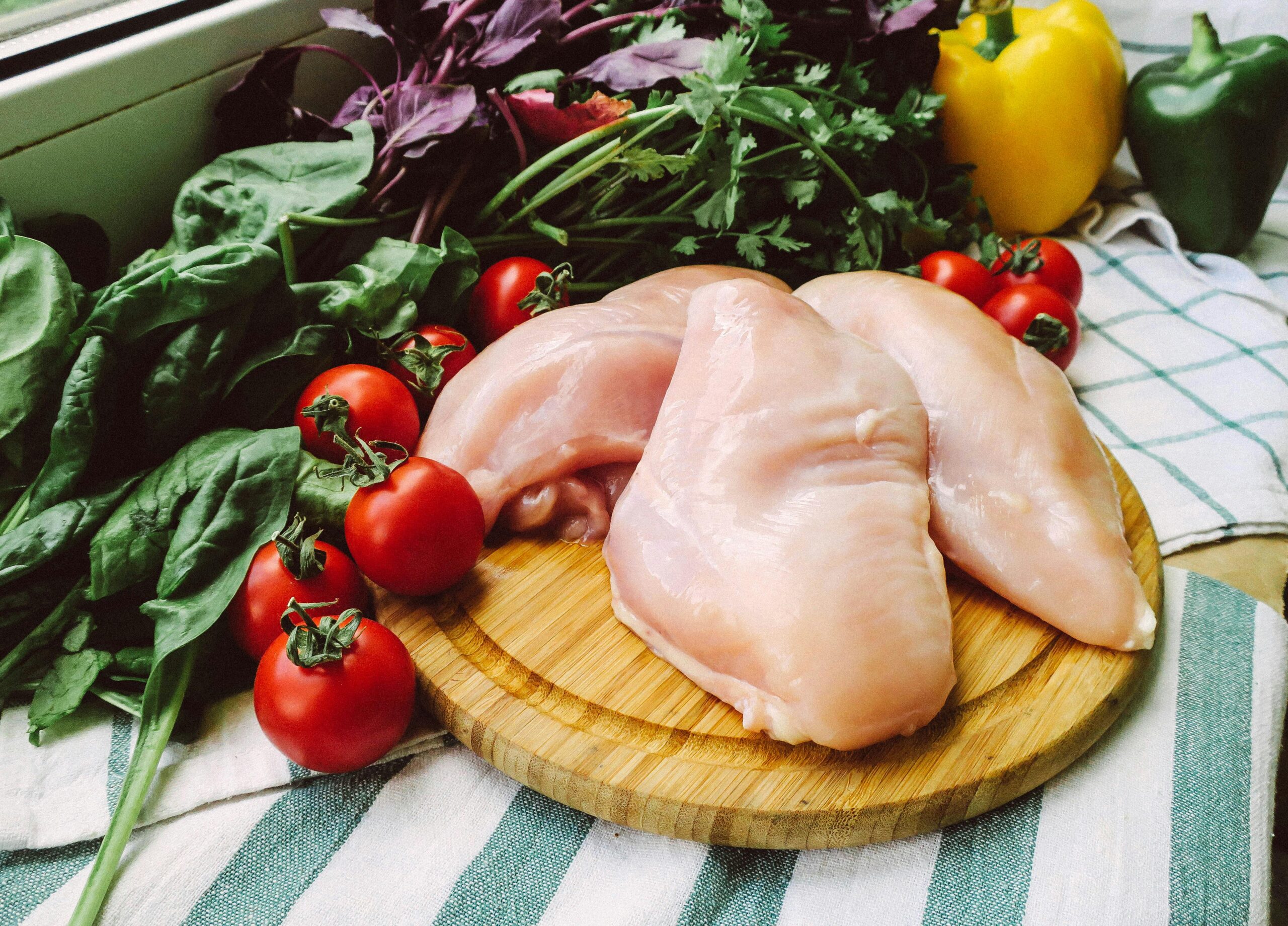 Raw chicken breasts on chopping board surrounded by fresh vegetables for cooking inspiration.